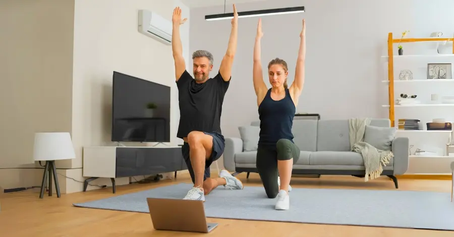 Casal treinando junto em casa, realizando um exercício com peso corporal para manter a forma sem necessidade de academia.