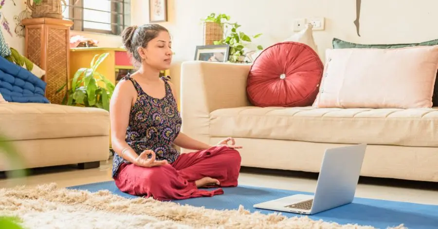 Mulher sentada em posição de lótus em um tapete de yoga em casa, meditando com um notebook aberto à sua frente, indicando uma prática online.